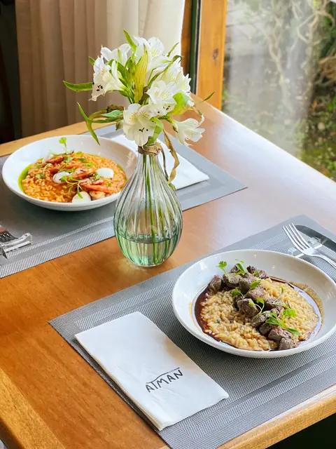 Mesa de madeira posta para o almoço, com pratos, flores e o jardim na janela